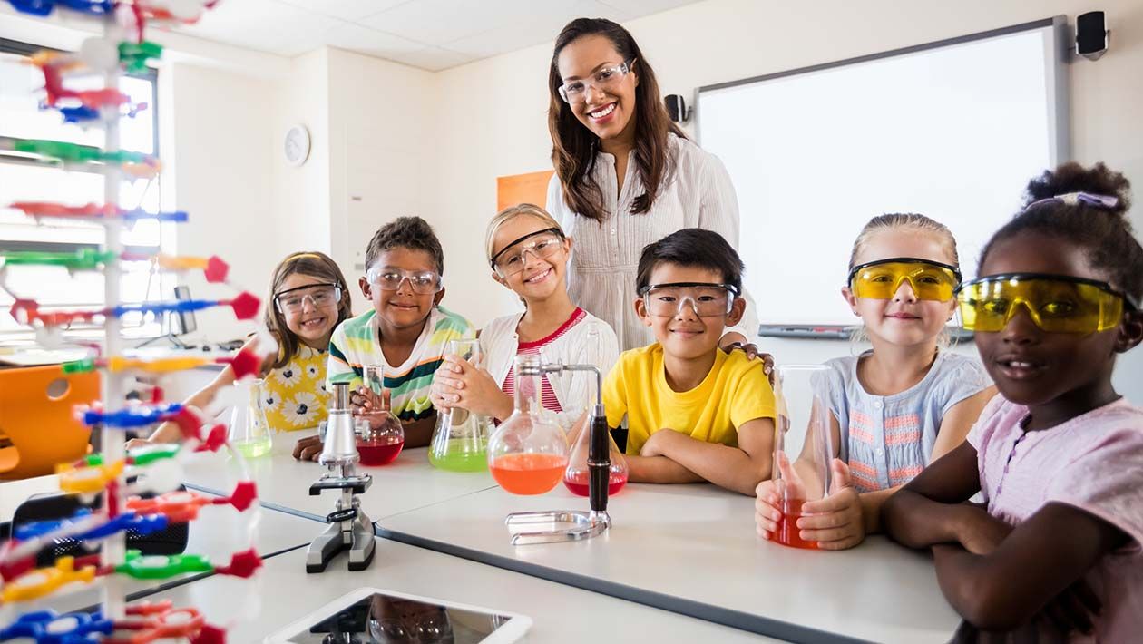 Students wearing safety goggles smiling at the table holding their beakers with multi colored liquid inside them.
