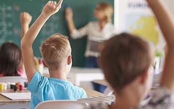 3 students in a classroom raising their hands while teacher writes on the blackboard.