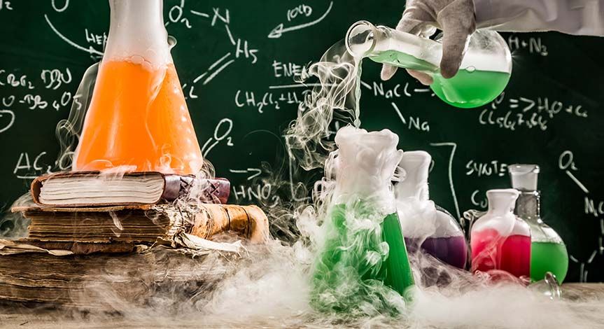 Hand pouring liquid into beaker on a desk.  Another beaker on a stack of books with smoke surrounding all beakers and table